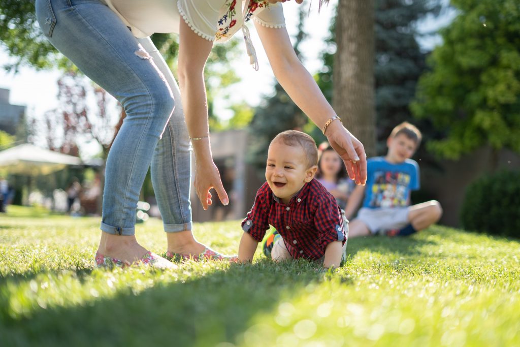 woman in white blouse and blue denim jeans helping a baby crawl on green grass
